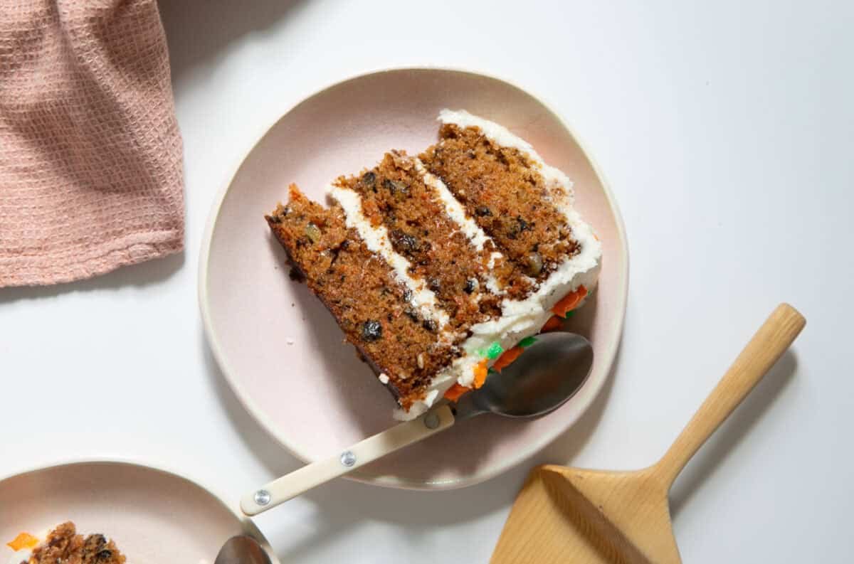 Slice the carrot cake on a plate with a spoon and cloth next to it.