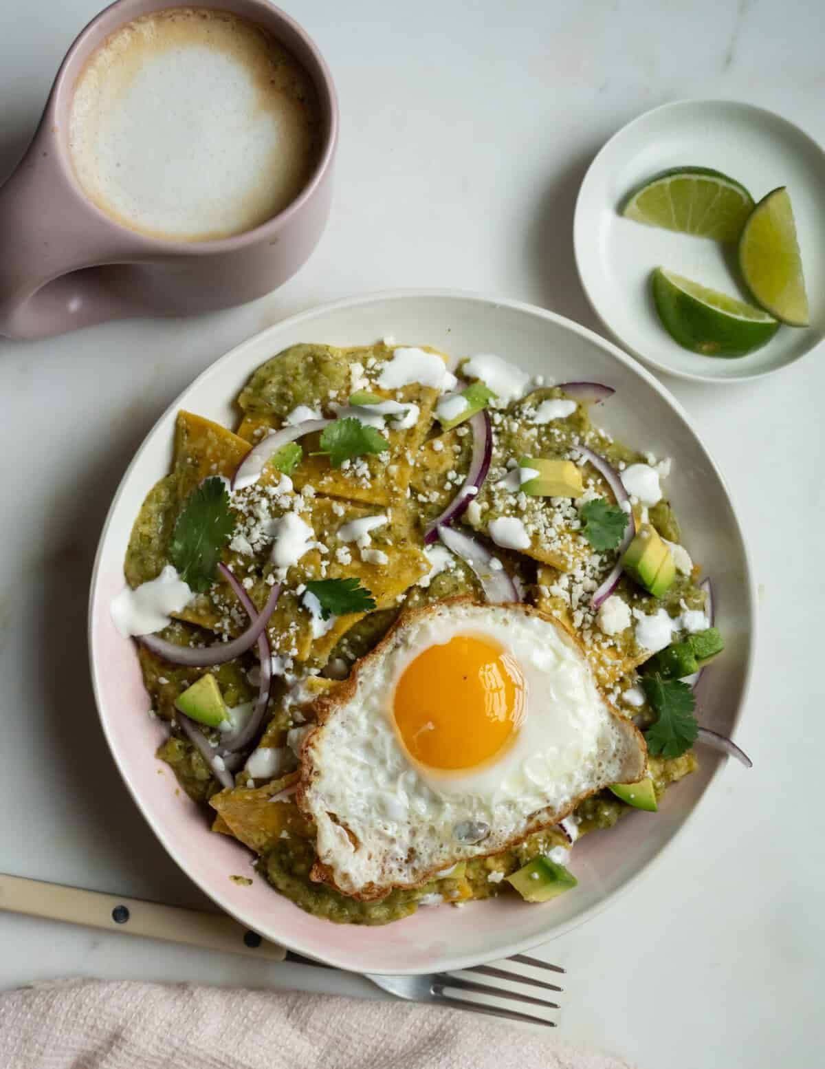 Chilaquiles are verdated in a bowl with a fried egg on top. Coffee and lime slices are on the side.