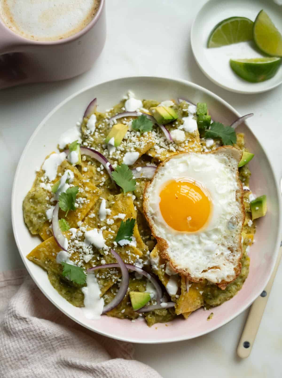 Chilaquiles are verdated in a bowl with a fried egg on top. Lime on the side with a cup of coffee.